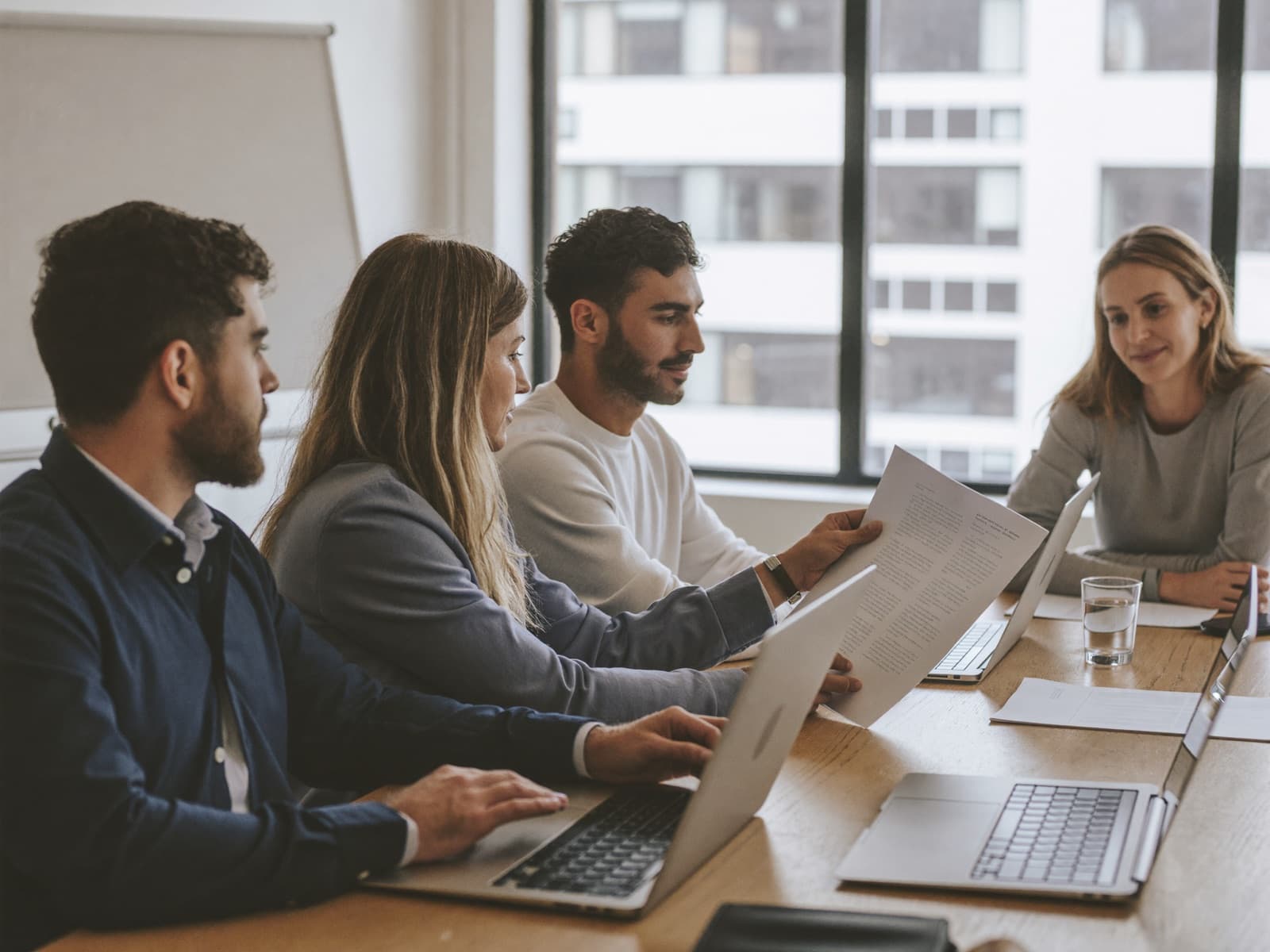 Three coworkers at a table, one holding up a printed draft while a colleague reads it over her shoulder.