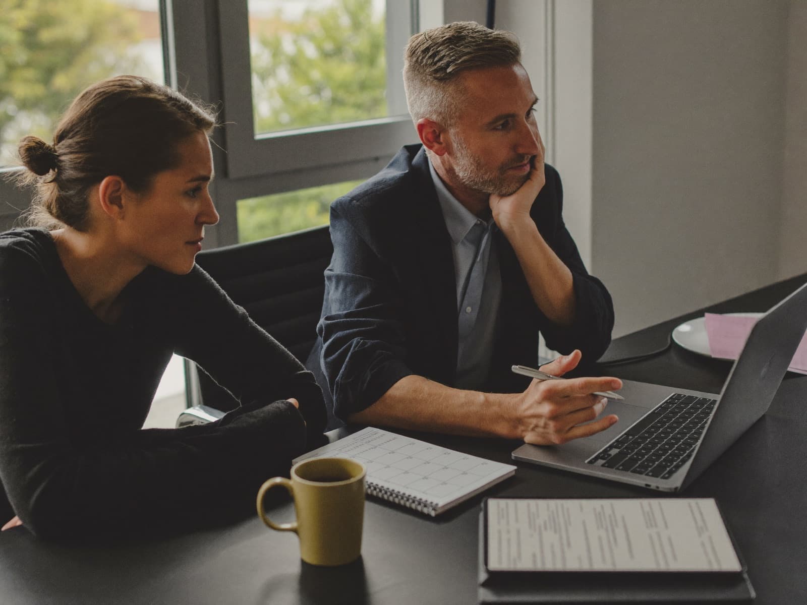 A coach sitting beside a business owner at his desk, both looking at his laptop.