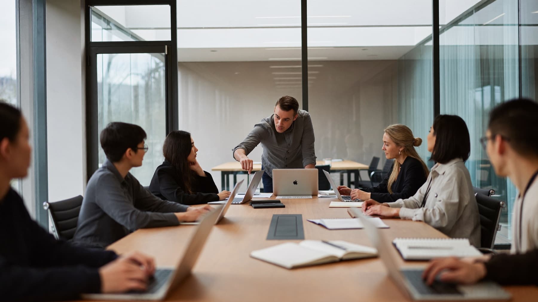 A coach leaning over a conference table to point at one person's laptop, while eight coworkers work on their own laptops.